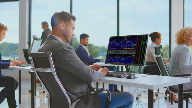 Finance professional working on a computer display showing multiple stock market charts and data in a vibrant, modern open-plan office setting with colleagues in the background