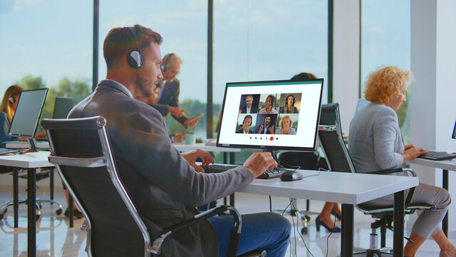 Businessman wearing a headset and talking during an online video call on a computer with remote colleagues, working in a contemporary open-plan office setting