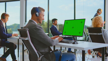 Customer service representative sitting at desk with headphones, typing on keyboard and looking at a computer monitor with a chroma key green screen in a busy call center office