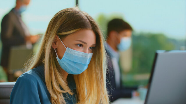 Young businesswoman wearing a protective face mask while concentrating on computer work in a contemporary open-plan office, highlighting new normal workplace safety protocols