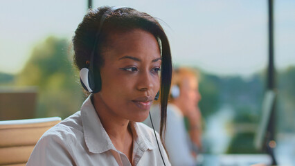 Black woman call center agent wearing a headset, focused on her computer screen while providing attentive customer support in a busy modern office environment