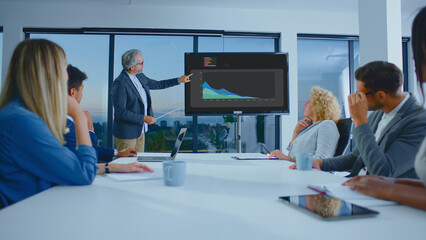 Professional business people actively participating in a corporate meeting, analyzing financial data presented on a screen by an experienced male speaker in a modern office boardroom