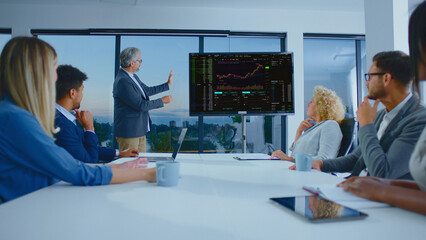 Group of diverse business professionals discussing financial data and analyzing stock market charts on a screen during a corporate presentation in a modern office meeting room