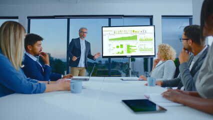 Confident businessman presenting financial data and charts on a large screen to a diverse team of colleagues during an important corporate meeting in a modern office boardroom