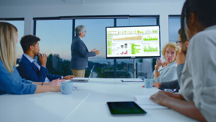Senior businessman presenting financial charts and data on a large screen to a diverse team in a modern conference room, collaborating on strategy, planning, and analysis