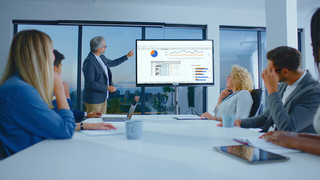 Senior businessman presenting financial data and charts on a large screen to a diverse team of colleagues participating in a corporate meeting in a modern office boardroom