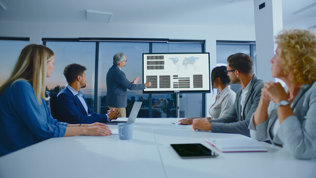 Experienced entrepreneur presenting financial data and world map analytics on a large screen to a diverse group of employees during a corporate meeting or seminar
