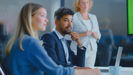 Diverse business professionals attending a corporate meeting in a modern office, a man in a suit thoughtfully listening while a woman presents near a green screen on a large display
