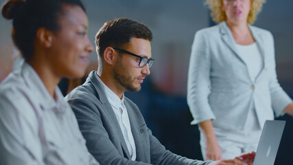 Diversified business team discusses project strategy and analyzes data on a laptop in a modern office while a colleague presents, highlighting collaboration and communication skills