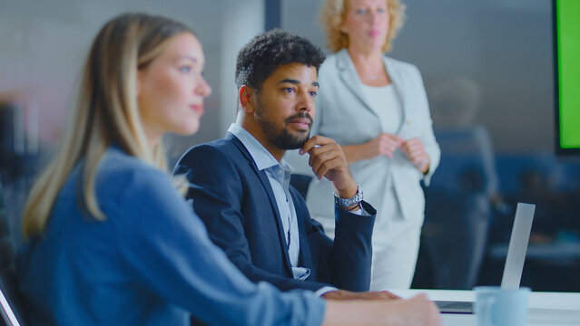 Diverse business professionals attending a corporate meeting in a modern office, a man in a suit thoughtfully listening while a woman presents near a green screen on a large display