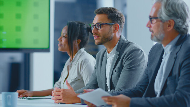 Diverse team of business professionals collaborating and discussing ideas during a corporate meeting in a modern office setup, featuring a large blank green screen for dynamic content