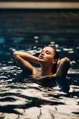 Beautiful young woman bathing in clear pool water, leaning back with eyes closed, illuminated by soft light, concept of relaxation and wellness.