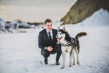 Stylish young man in a formal suit lovingly crouches with his happy Siberian Husky dog in a snowy winter landscape.