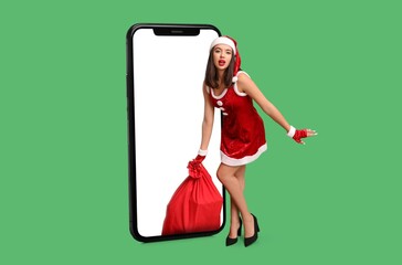 Sexy young woman in Santa costume and big smartphone on green background