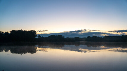 Obraz premium Wide, tranquil landscape view of a lake reflecting the soft blue and yellow hues of the sky at dawn during the blue hour. Naplál lake, Budapest.