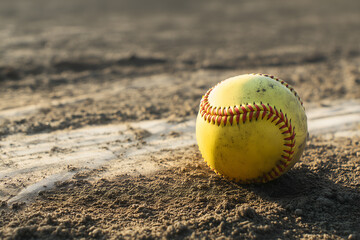 Yellow softball with red stitching lying on dusty infield near baseline, minimalist sports still life.