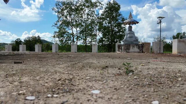 The images depict a wide open outdoor scene featuring a paved area with some patches of dirt, leading towards a large statue of a seated figure holding an umbrella, situated on a raised platform