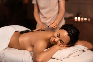 Young African-American woman getting massage with stones in dark spa salon