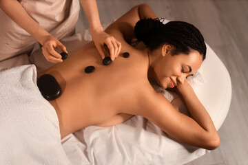 Young African-American woman getting massage with stones in dark spa salon