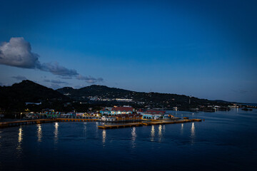 Scenic blue hour view of Roatán island in Honduras, featuring illuminated waterfront buildings and calm sea reflections under a clear evening sky. Hills in the background and warm lights.