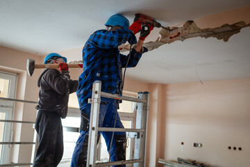 Two construction workers on scaffolding demolishes a wall in a building with a hammer drill.