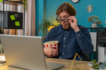 Caucasian businessman freelancer taking break from work wearing 3D glasses eating popcorn and watching movie on laptop. Laughing young man sitting at home office desk having snacks during break.