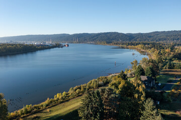 Aerial drone picture of the Columbia River from Sauvie Island in Portland Oregon, showing calm blue water, surrounding trees, and forest park, on a clear sunny fall morning during the autumn season
