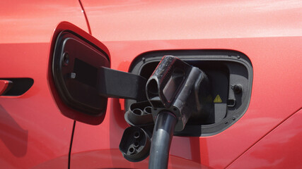 Close-up of a vibrant red electric car being recharged at a public station, showcasing the handle and cable in the port, symbolizing clean energy, zero-emission travel, and modern electric mobility