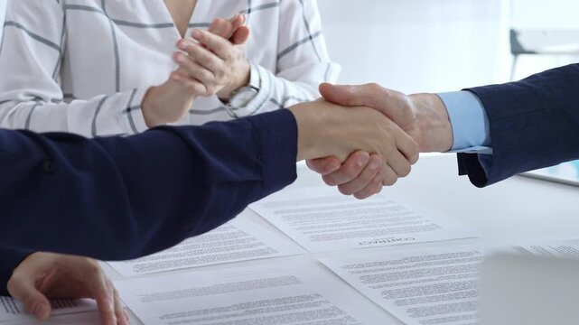 Business people are shaking hands and applauding, finalizing a deal while reviewing financial documents, graphs, calculator and charts on a desk in a modern office environment