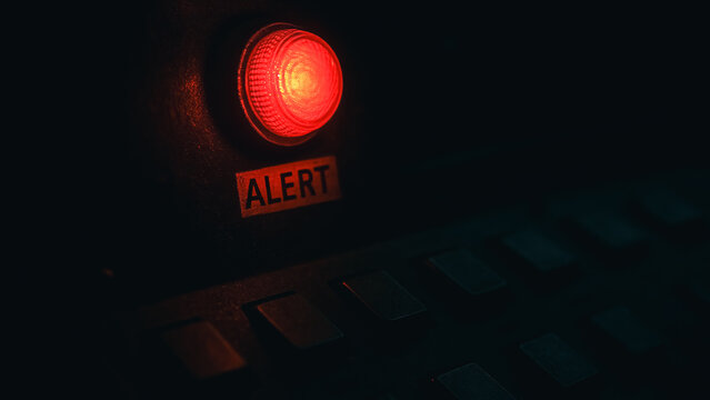 A close-up shot of an 'ALERT' button illuminates, casting a stark, red glow in a dim environment. The illuminated button becomes the central point of focus, drawing the viewer's eye.