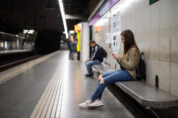 Young woman uses smartphone in a subway station as she waits for her train, connecting with friends and family, or catching up on social media during her commute
