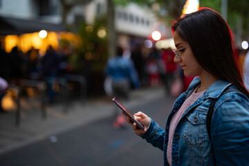 Trendy young woman checking her phone while strolling through the city at dusk, connecting with friends and family, enjoying modern life with easy communication, stylish and relaxed