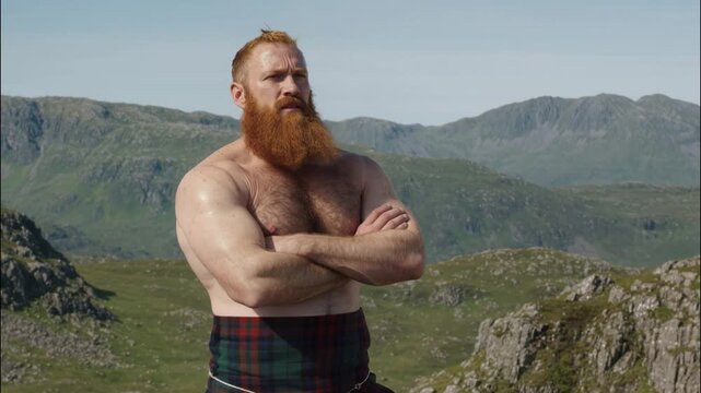 Strong red-bearded man wearing a traditional Scottish kilt standing confidently with folded arms in a scenic mountain landscape under a clear sky.
