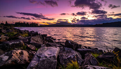 Purple Hued Sunset Over Calm Lake Waters with Rocky Shoreline in Foreground Scenery with Silhouette of Trees on Horizon Reflective Water Serene Landscape