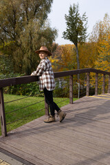 Fototapeta premium Caucasian woman with long hair stands on a on the wooden bridge surrounded by autumn trees. She wears a plaid shirt and a wide-brimmed hat.