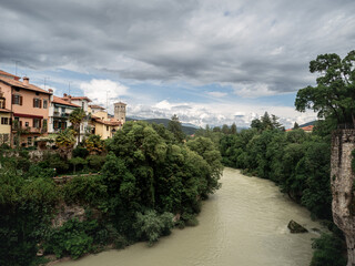Fototapeta premium View of the River Natisone winding through lush greenery and colorful houses in Cividale del Friuli, Italy. Overcast skies create a dramatic atmosphere above the peaceful riverside town.