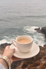 Cup of hot coffee overlooking the sea and coastal rocks. Serene morning vibe, ideal for vacation, mindfulness, and nature concepts.