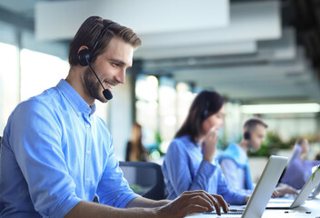 Smiling male call-center operator with headphones sitting at modern office with collegues on the backgroung, consulting online.