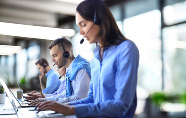 Smiling male call-center operator with headphones sitting at modern office with collegues on the backgroung, consulting online.