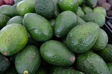 The image shows a large pile of avocados displayed on a market stand. The avocados are dark green with a slightly bumpy texture, typical of Hass or similar varieties.