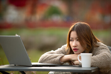 A young Asian woman, dressed in a cozy fleece, appears deeply contemplative or perhaps a bit bored, leaning on her arms at an outdoor table with a laptop and a white coffee mug. The soft, blurred back