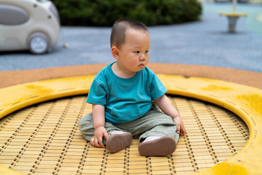 A young Asian child in a green short-sleeved shirt, not yet able to walk, sat on the trampoline at the playground, looking utterly bewildered.