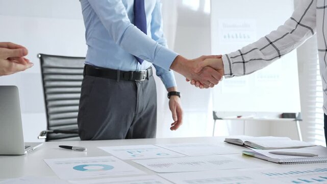 Business partners shaking hands in office with financial charts, laptop and reports on table, symbolizing success, teamwork, agreement, collaboration and corporate growth. Businesspeople handshake