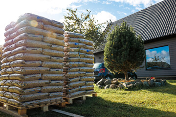 Two stacks of bagged materials on pallets near a modern building with a dark roof, a landscaped area with a tree, and a partially visible parked car.