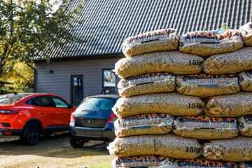 A stack of 15kg wood pellet bags in sunlight, with a gray wooden house, sloped metal roof, red and dark colored cars, and green trees in view.