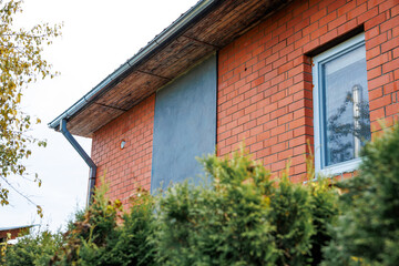 Part of a red brick building with a slanted roof, a reflective window, and green shrubs in the foreground. A tree with yellowing leaves is on the left.