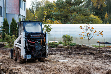A small construction vehicle with exposed engine parts sits on muddy ground near a residential building, with a greenhouse and young trees nearby.