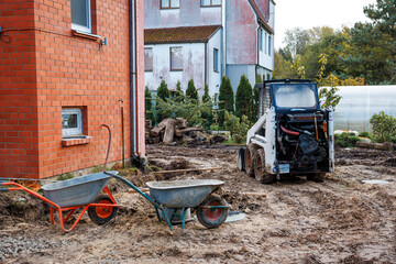 A white skid steer loader on muddy ground near a brick wall with red and blue wheelbarrows. Residential buildings and a greenhouse are visible nearby.