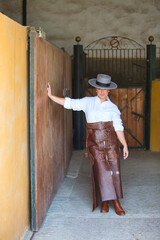 A young, attractive woman dressed in riding clothes and a traditional Spanish hat is leaning against a wall with one hand. She is looking straight ahead and appears happy.