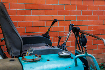 Detailed view of a blue tractor's control area, featuring levers and a seat. The tractor is set against a red brick wall, showing signs of regular use.